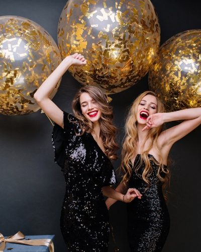 Happy party moments of two fashionable funny young women on black background. Luxury black dress, red lips, long curly hair, brightful mood, having fun, big balloons with golden tinsels.